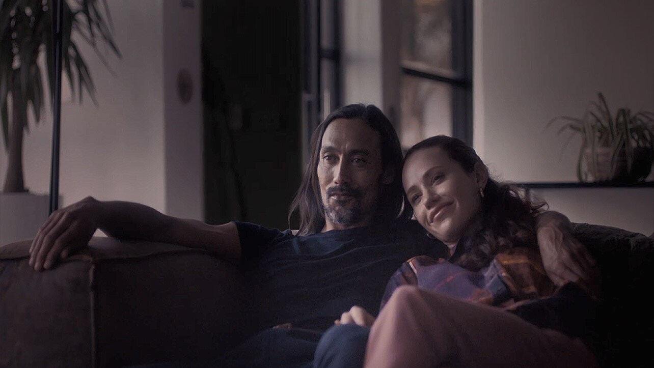 Man with his arms around a woman on a couch while looking forward (play the video)