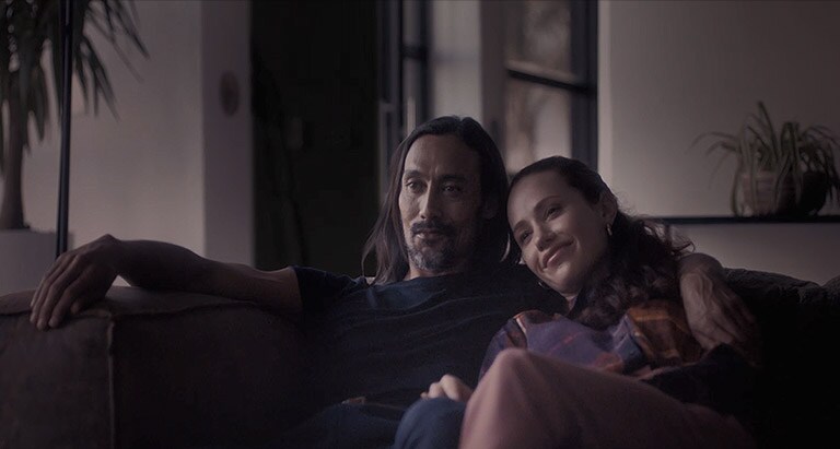 Man with his arms around a woman on a couch while looking forward (play the video)