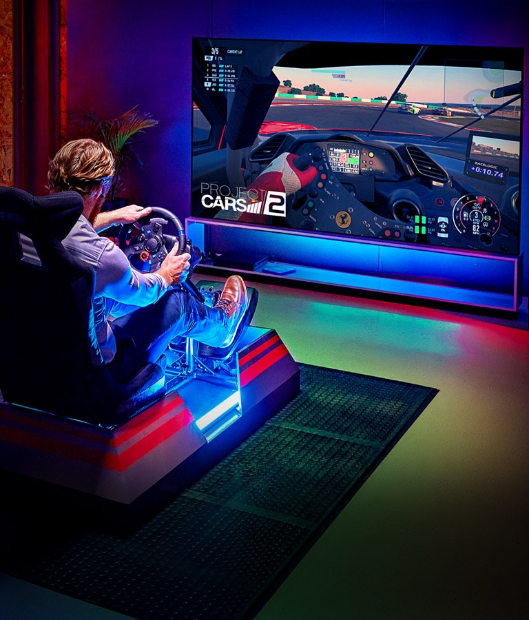 Man in a dark room sitting on a racing game seat in front of a large TV playing a racing game (scroll down the page)