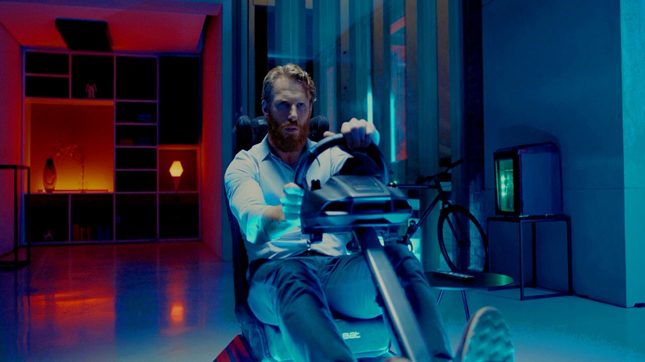 Back shot of a  man sitting in the room on a racing game seat playing a game in front of a large screen TV (play the video)
