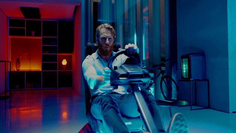 Back shot of a  man sitting in the room on a racing game seat playing a game in front of a large screen TV (play the video)