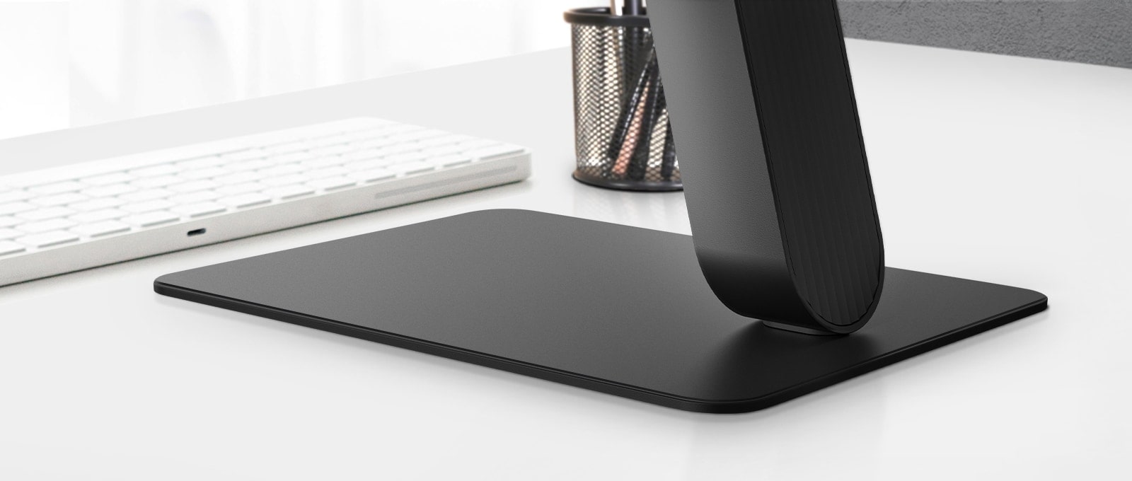 A close-up view of a sleek black monitor stand with a flat rectangular base, placed on a white desk beside a white keyboard and pen holder. The minimalist design emphasizes stability and elegance, blending seamlessly into a modern workspace.