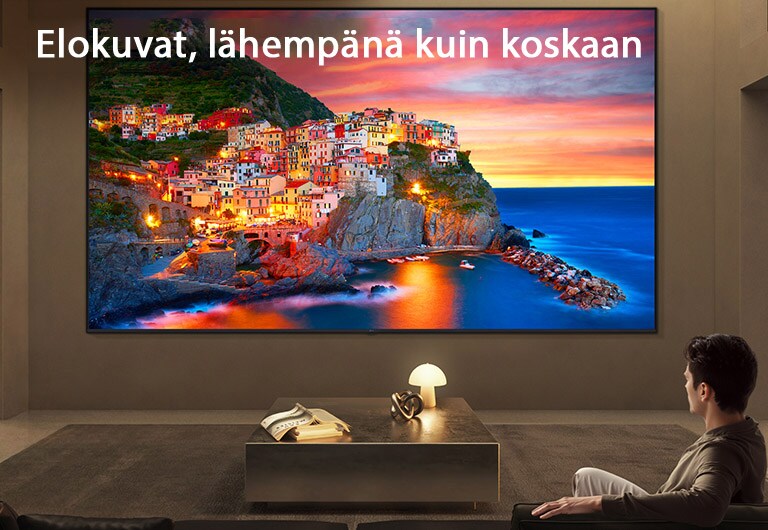 LG Ultra Big TV on seinäasennettuna tilavassa olohuoneessa, jossa laaja näyttö esittää eloisan rannikkokylän auringonlaskun aikaan, havainnollistaen kuinka suurempi näyttö tarjoaa mukaansatempaavan ja elokuvamaisen katselukokemuksen.