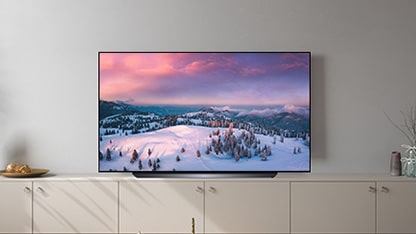 LG OLED CS is sitting on top of cabinets in a softly lit room with a snowy landscape on-screen.