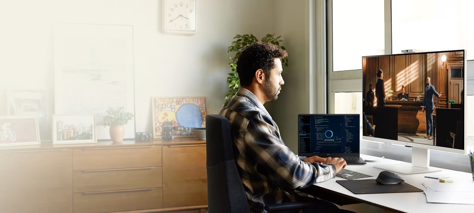 personne travaillant à un bureau avec un moniteur LG et un ordinateur portable. Le moniteur affiche une scène d’une vidéo ou d’un film, tandis que l’ordinateur portable affiche du code ou des données. L’espace de travail est lumineux et organisé, avec des plantes, des œuvres d’art et une fenêtre qui apporte de la lumière naturelle.