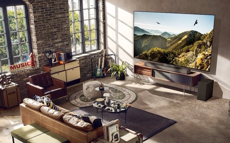 Un salon loft spacieux et élégant avec une grande TV murale affichant une vue sur les montagnes. Le décor inclut des murs en briques, de grandes fenêtres industrielles, un canapé en cuir et des instruments de musique, créant une ambiance chaleureuse et artistique.