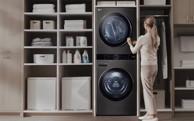 Une femme utilise un lave-linge et sèche-linge noirs empilés dans une buanderie élégante et bien organisée, avec des étagères et des armoires en bois remplies de produits de lessive et de serviettes propres.
