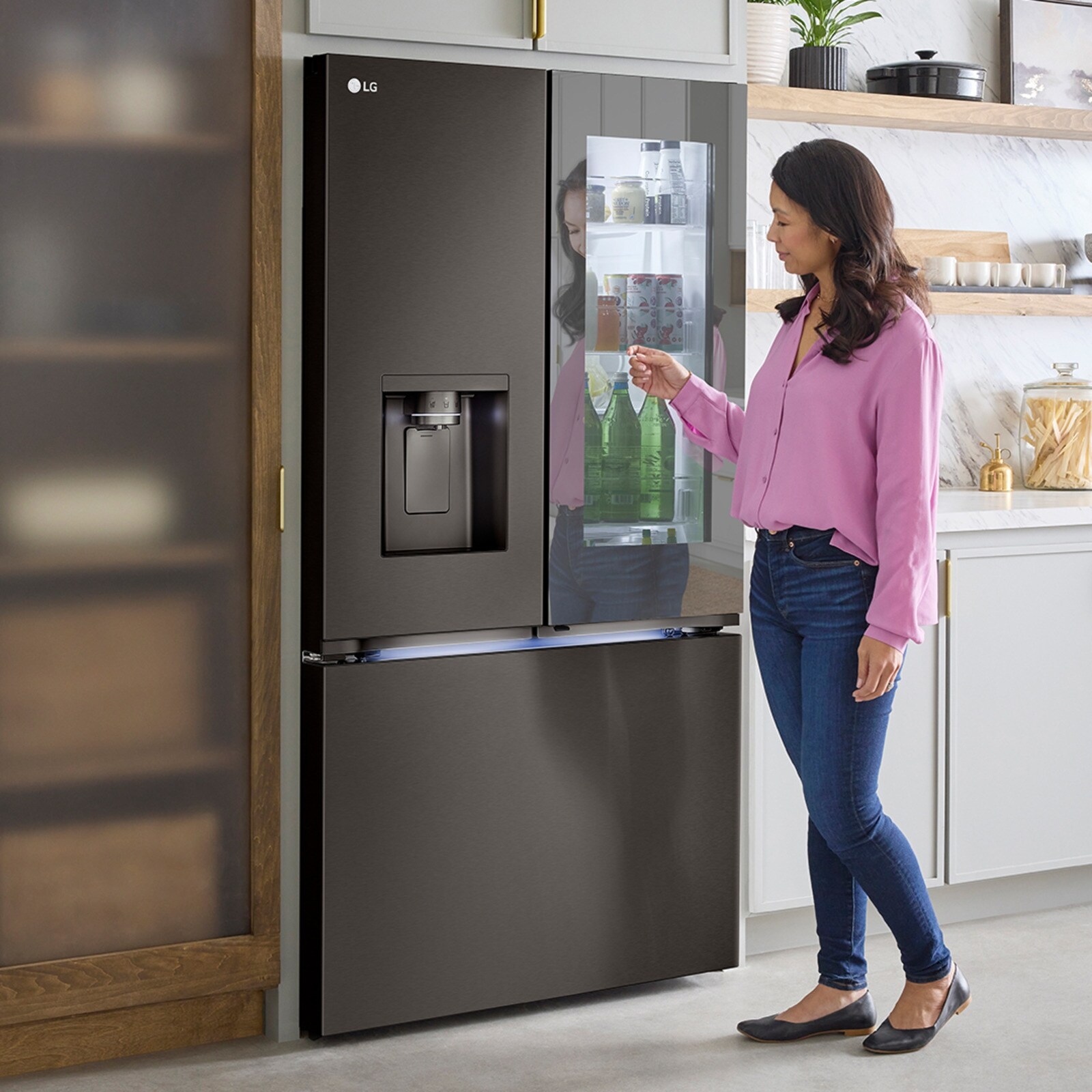 Une femme interagit avec un réfrigérateur moderne en acier inoxydable LG à portes françaises, doté d’un design « Door-in-Door ». Elle accède au compartiment facile d’accès où sont stockés les boissons et les articles fréquemment utilisés. Le réfrigérateur est également équipé d’un distributeur d’eau et de glaçons intégré dans la porte gauche.