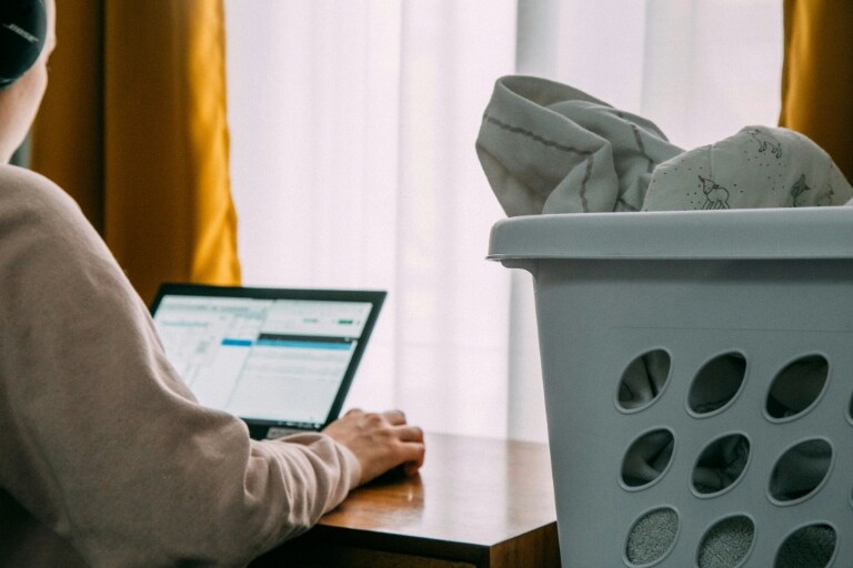 Une personne utilise un ordinateur portable à côté d’une fenêtre avec des rideaux jaunes, tandis qu’un panier à linge rempli de vêtements se trouve au premier plan.