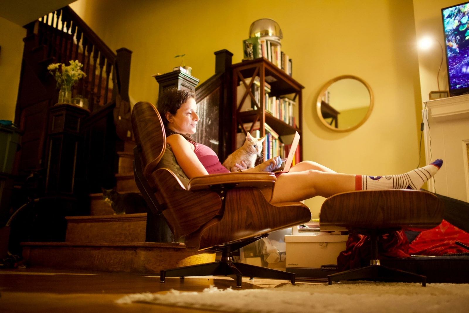 Femme écrivant sur son ordinateur portable, installée dans un fauteuil inclinable dans son salon, avec un éclairage tamisé et une télévision allumée en arrière-plan.