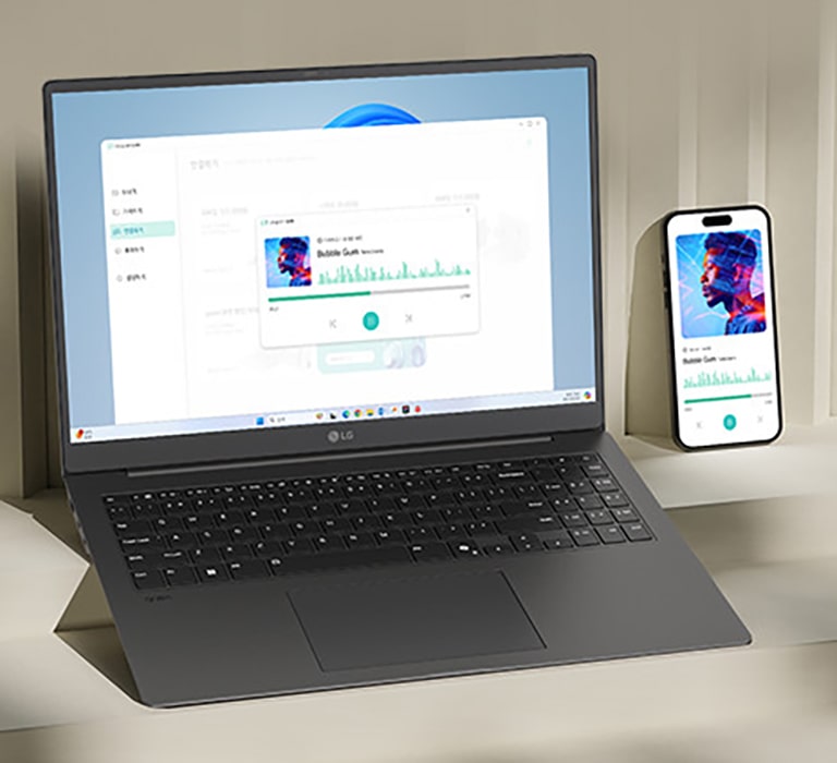 A laptop and a smartphone are placed on a desk, both displaying the same music playback interface, indicating synchronized functionality. The setup highlights seamless integration between devices for multimedia activities.	