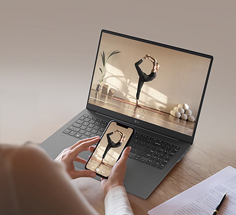 The image features a person holding a smartphone in front of a gray laptop, both displaying the same video of a woman performing a yoga pose. The scene highlights seamless screen mirroring or content sharing between devices.	