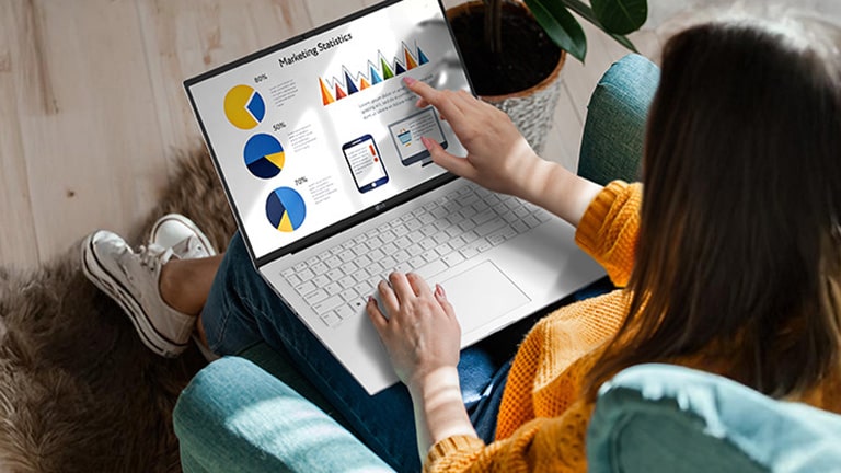 The image shows a person using a laptop while seated comfortably, interacting with the touchscreen to navigate charts and graphs. An animation demonstrates the anti-glare screen by showing no light reflection even when sunlight shines directly onto the display.	