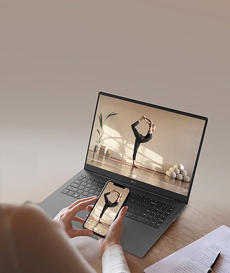 The image features a person holding a smartphone in front of a gray laptop, both displaying the same video of a woman performing a yoga pose. The scene highlights seamless screen mirroring or content sharing between devices.	