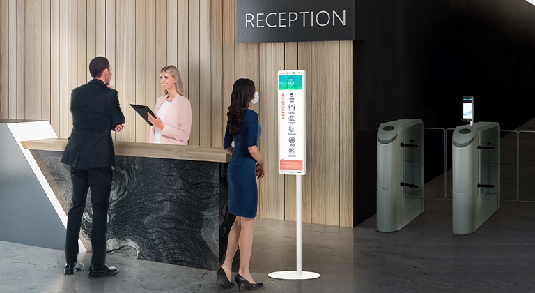 The 29" Thermal sensing terminal is placed near the reception desk. And The employee with a mask stands in front of the terminal to check her fever .