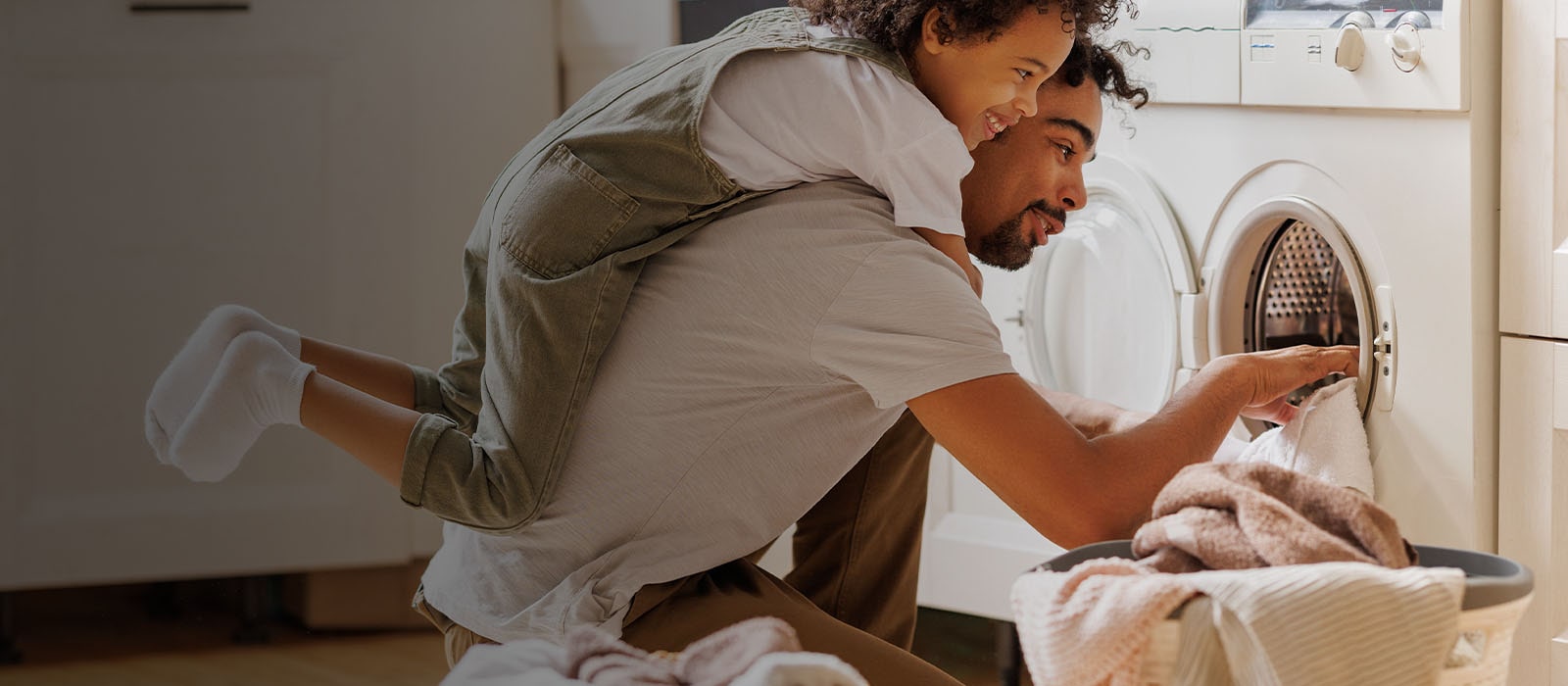 The picture shows a father and son doing laundry together, the son is giving a hug to his dad from behind, happily smiling. 