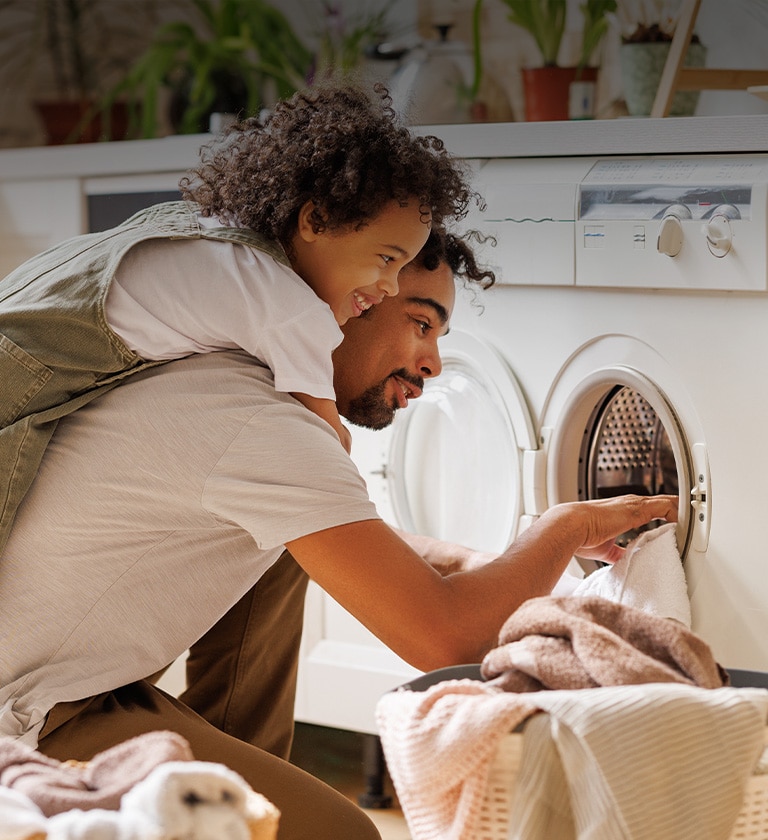 The picture shows a father and son doing laundry together, the son is giving a hug to his dad from behind, happily smiling. 