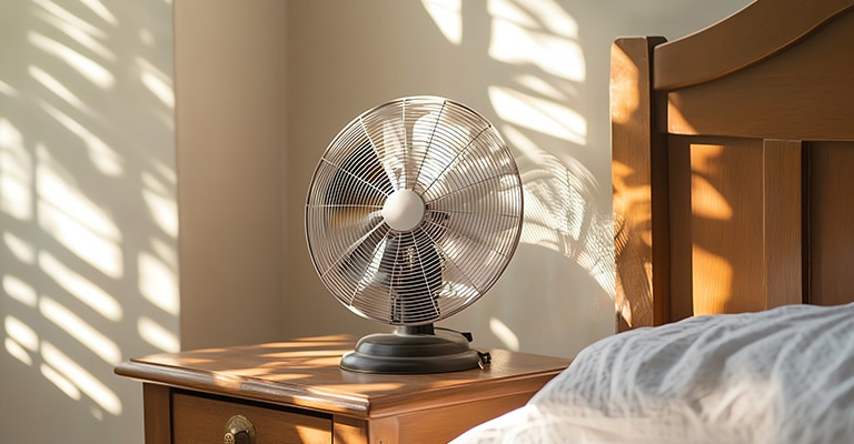 An electric fan set on a wooden table beside a bed.