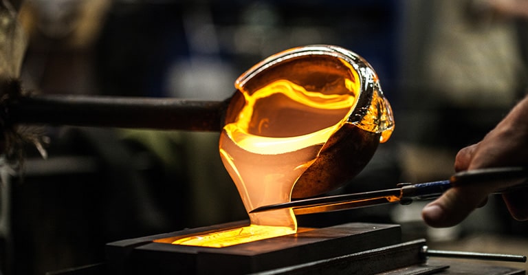 A close-up of a glassblowing process, focusing on a large blob of molten, glowing glass being worked.
