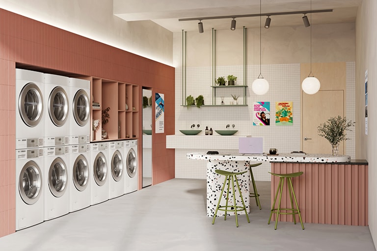 A multi-housing with rows of lg washing machines and dryers. The interior features a red accent wall, potted plants, and a seating area with a marble counter and colorful stools.