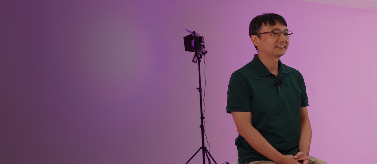 LG CAE Team Member wearing green short-sleeved shirt is sitting on a chair with pink background.