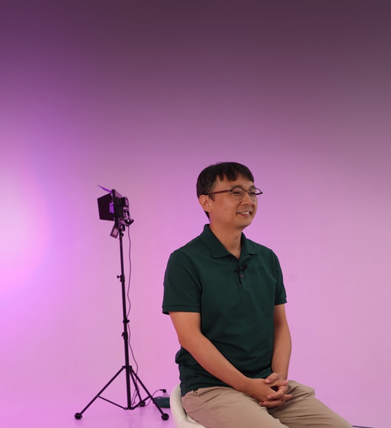 LG CAE Team Member wearing green short-sleeved shirt is sitting on a chair with pink background.