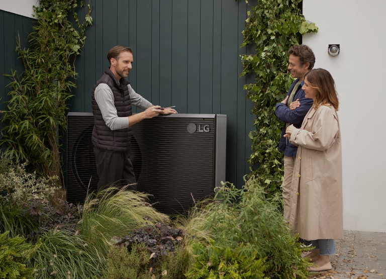 Man demonstrating LG Therma V outdoor heat pump, speaking to a woman standing next to another person in a garden.