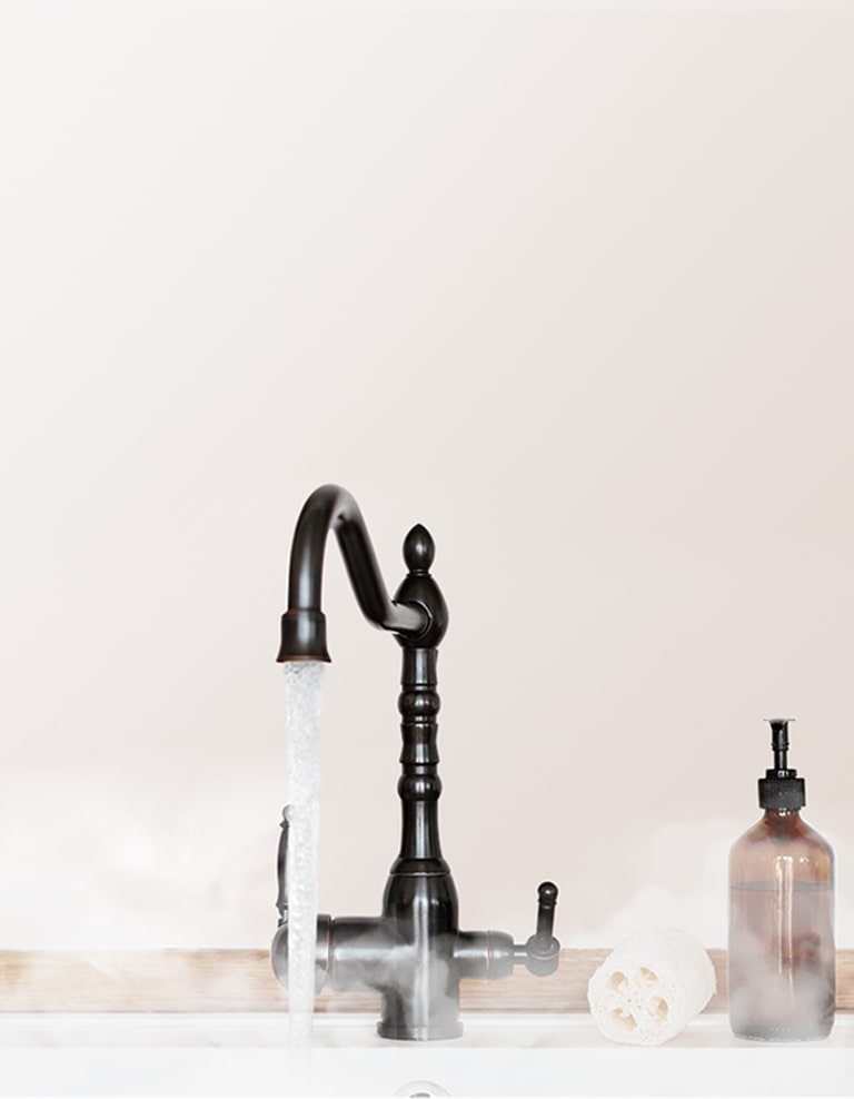 Steamy hot water pours from a black faucet into a white sink below, with a scrub and brown soap dispenser waiting at the faucet's right side.
