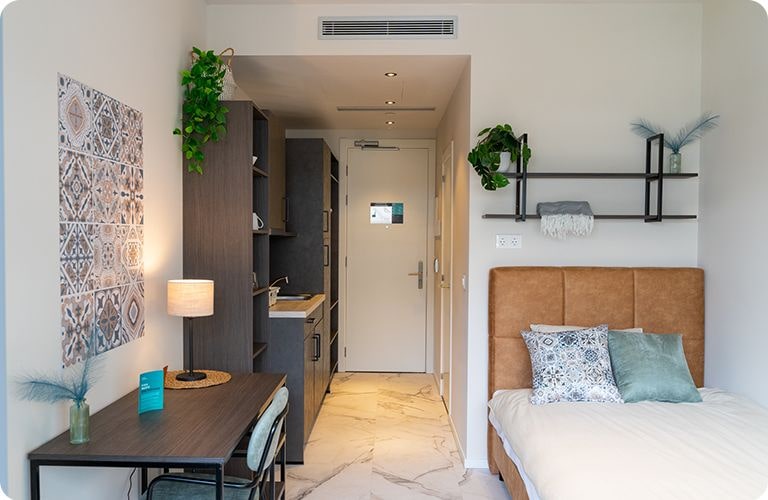 A compact room featuring a bed, a desk, and a small kitchen area, designed for efficient living and comfort.