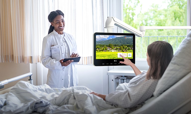In a hospital room, a patient is lying in a bed and touching an arm TV screen.
