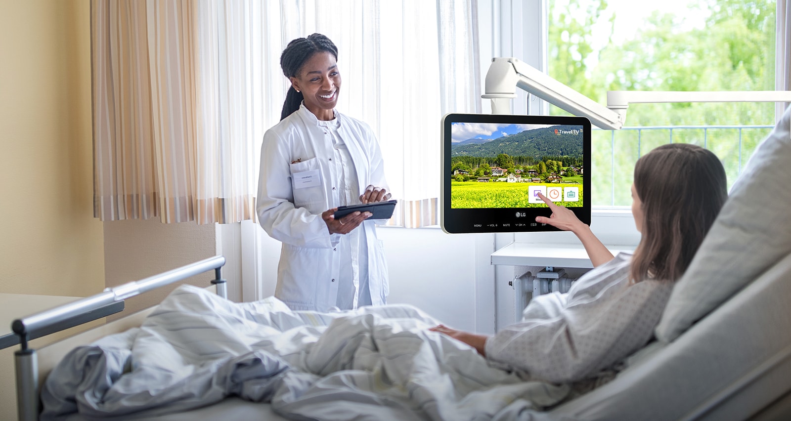 In a hospital room, a patient is lying in a bed and touching an arm TV screen.