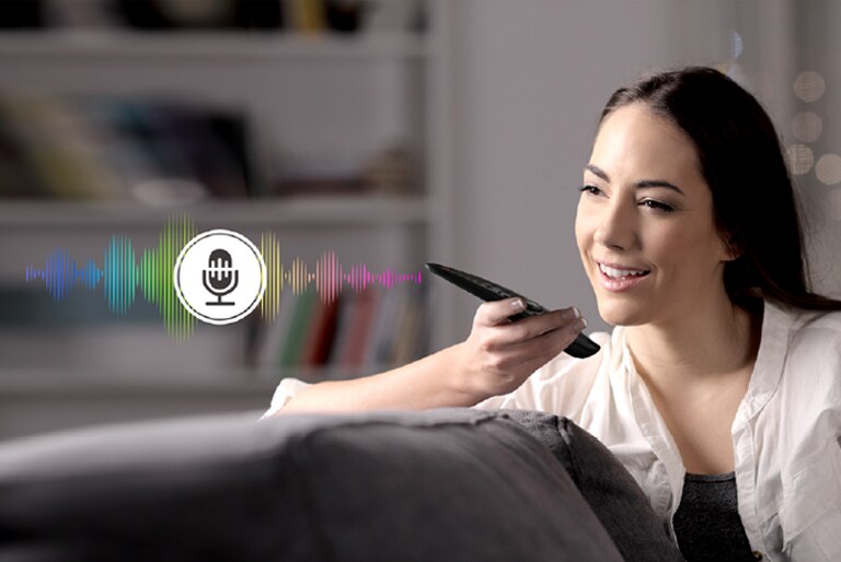 A woman is controlling TV by speaking to a remote control which has voice recognition.