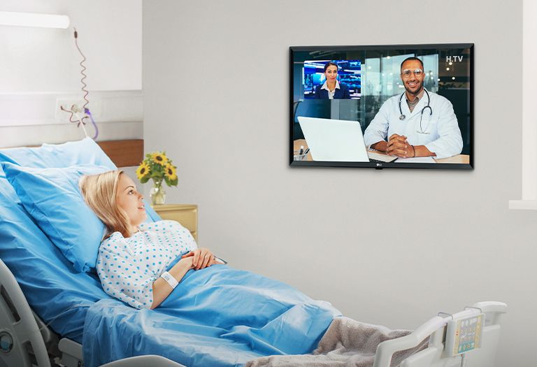 The patient is lying in the hospital room’s bed and watchiccng the TV hanging on the wall.
