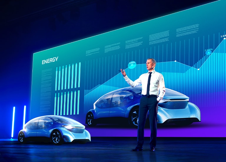 A man is giving a presentation in front of a large LED screen. The screen displays automobile-related content with clear visibility.