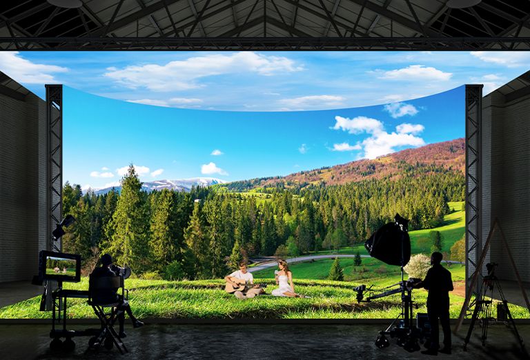 A virtual production studio with curved LED walls and ceiling and floor LEDs is filming images of a forest and field landscape.