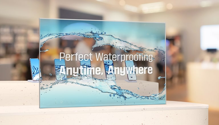 Transparent OLED Signage is installed in front of a table upon which cell phones are displayed in a cell phone store. The Transparent OLED Signage screen is showing an advertisement, and the cell phones displayed behind the screen are also clearly visible.
