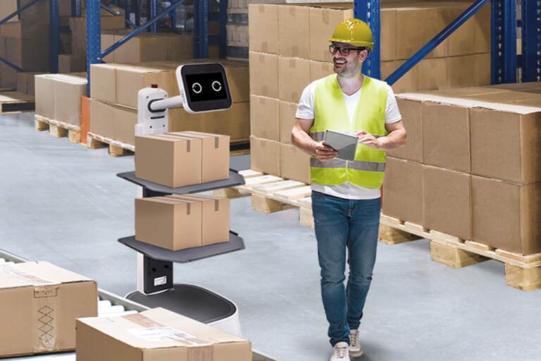 LG CLOi CarryBot is carrying two small boxes on each tray working right near the conveyor belt and a man is walking towards with a smile.	