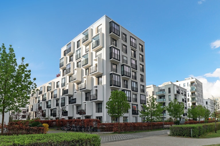 The image shows a modern apartment building with unique, staggered balconies. The area is surrounded by greenery, trees, and a clear blue sky, creating a clean and contemporary atmosphere.