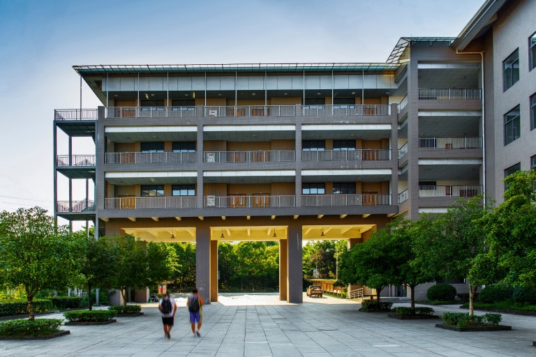 Modern dormitory building with multiple floors, large open passageway, balconies, and green trees surrounding the courtyard.