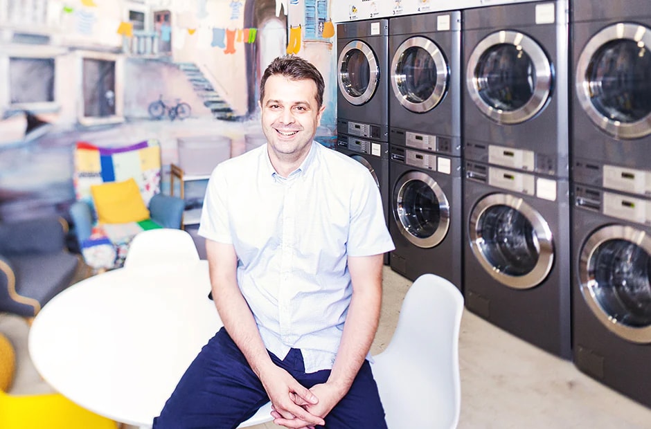 A man sits smiling in a colorful laundromat, with LG commercial laundry machines stacked in rows behind him.