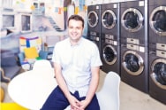 A man sits smiling in a colorful laundromat, with LG commercial laundry machines stacked in rows behind him.