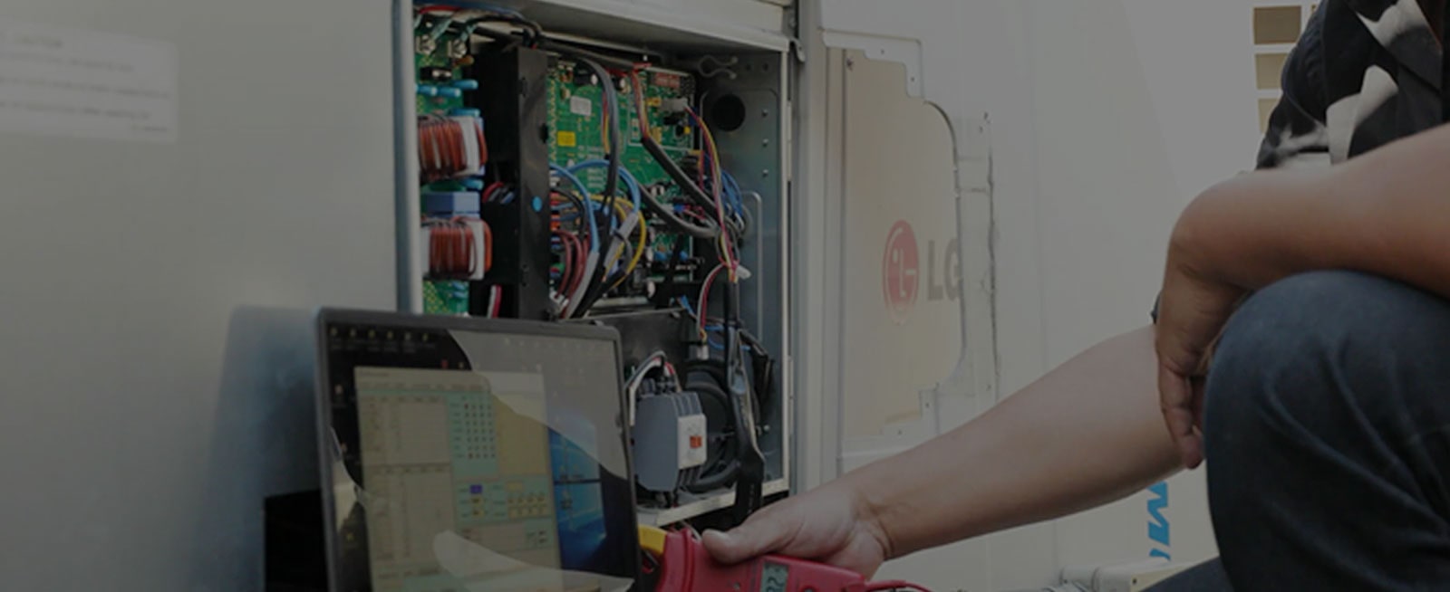 A person is using a red rectangular tool connected to a laptop, diagnosing an open LG outdoor unit, its internal wiring fully exposed.