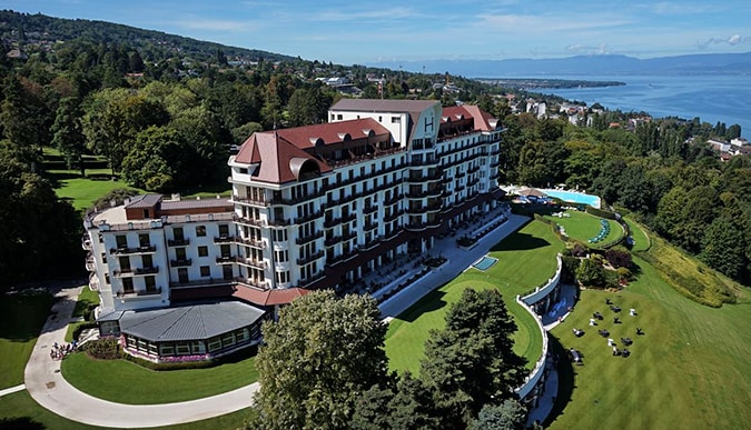 A birds-eye view of the Hotel Royal, Evian Resort situated on a hillside overlooking trees and a body of water.