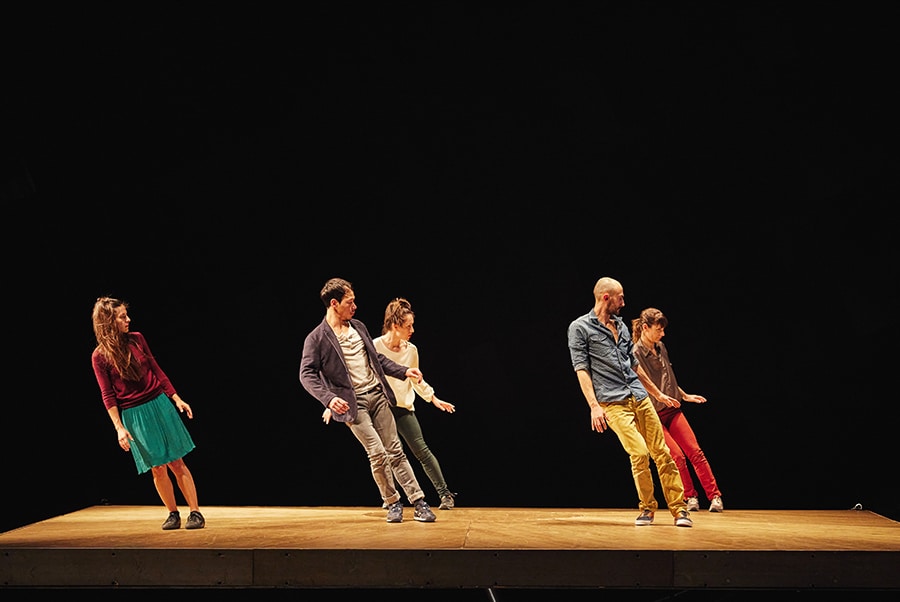 A photo of a handful of male and female dancers standing obliquely on a visible surface, with their glance uniformly cast downwards.