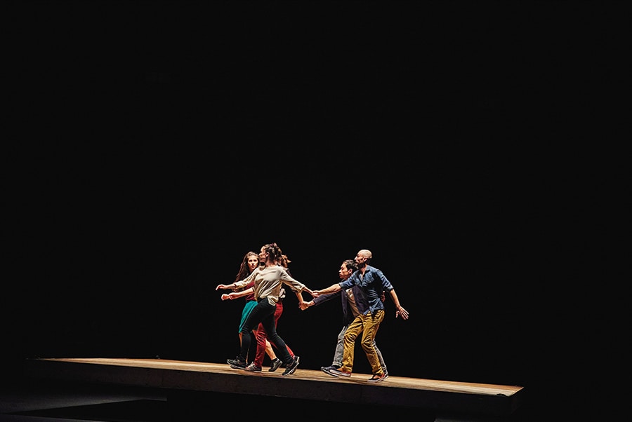 A photo of the group of dancers holding each other's hands as they walk on a slanted surface.