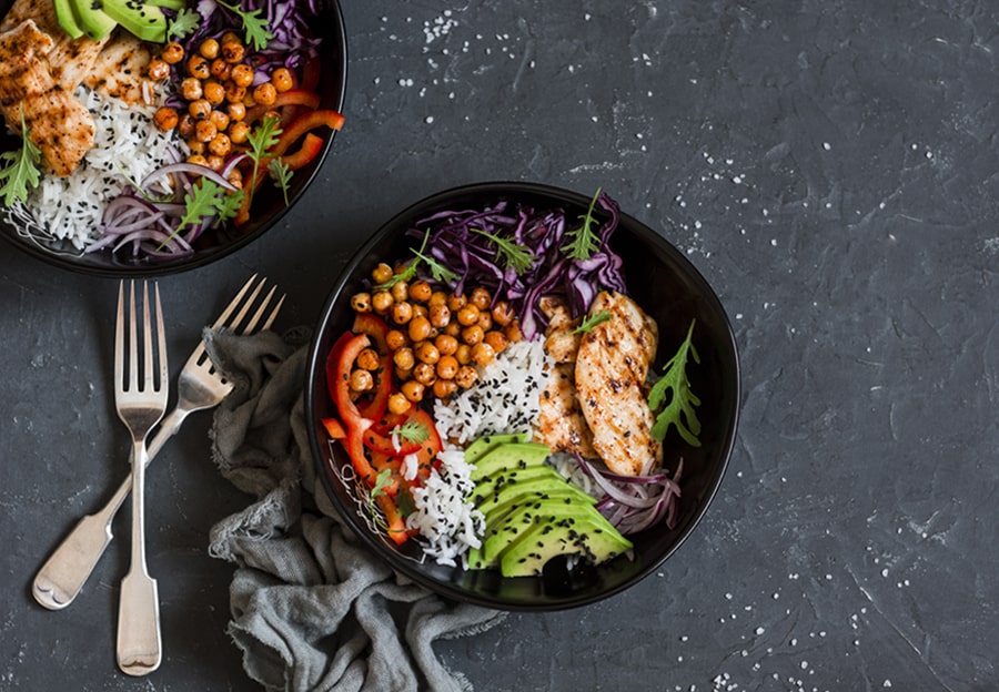 Two bowls of hearty chicken, chickpea, and avocado salad in black bowls on a black stone surface with two crossed forks