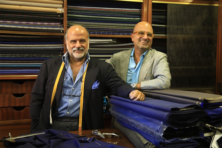 Two men stand in front of shelves containing bolts of fabric.