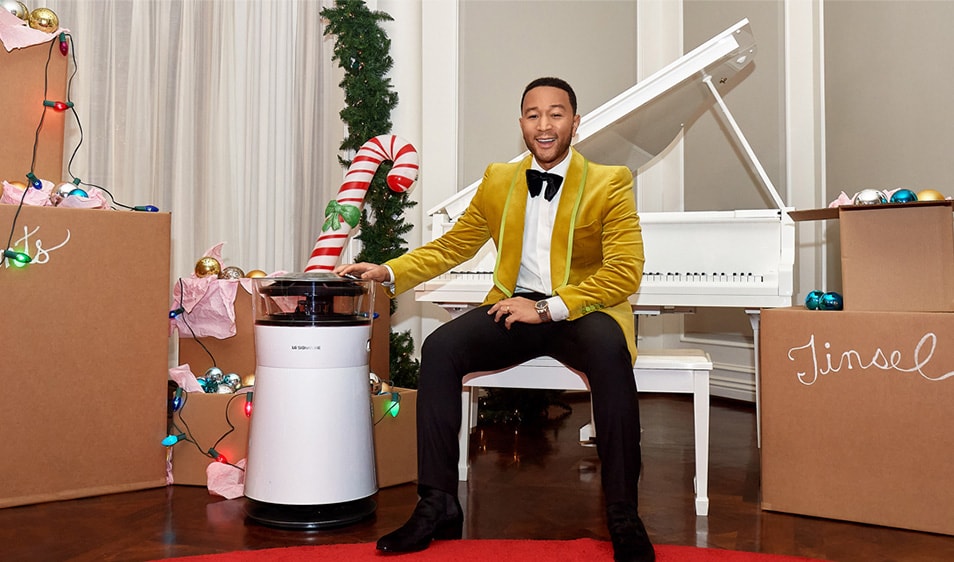 John Legend sitting at a white piano, resting his hand on an air purifier