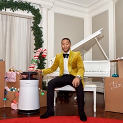 John Legend sitting at a white piano, resting his hand on an air purifier 