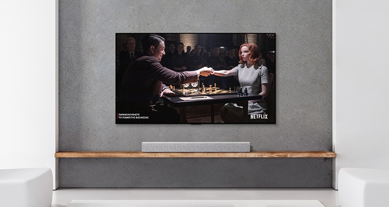 Ένα soundbar και μια τηλεόραση σε ένα σαλόνι με λευκή απόχρωση. Στην οθόνη της τηλεόρασης φαίνεται ένας άντρας και μια γυναίκα που παίζουν σκάκι.